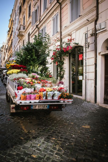 A delivery truck parked on a cobblestone street in front of a European-style building with pastel-colored facades and shuttered windows. The truck's flatbed is loaded with an array of fresh flowers, i