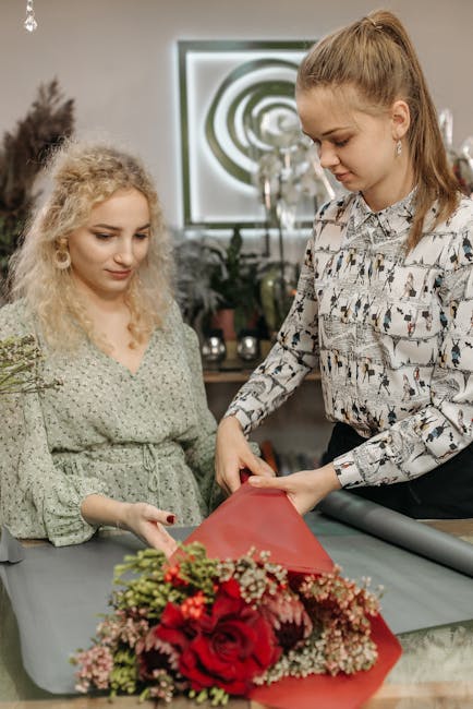 A floral arrangement being prepared by two women in a well-lit indoor space, possibly a florist shop or workshop. The bouquet features vibrant red roses, pink baby's breath, and assorted greenery, wra