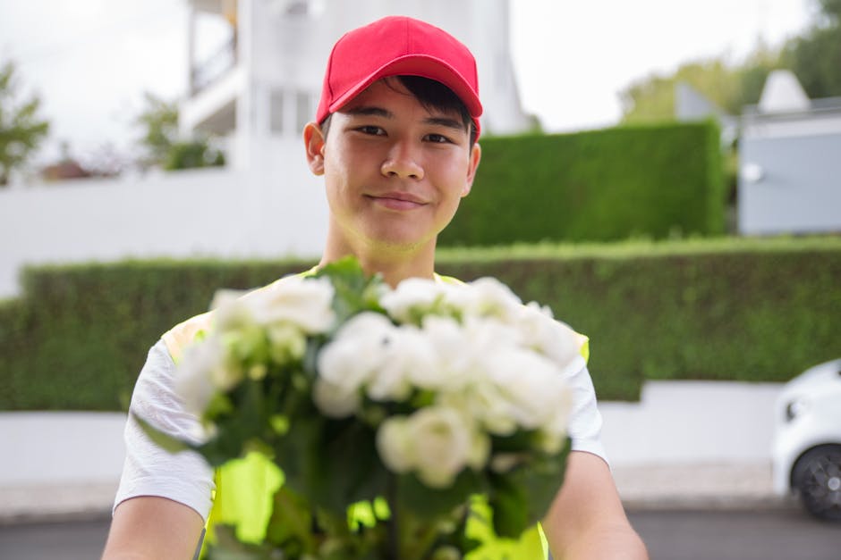 A young delivery person with a friendly expression, wearing a red cap and a high-visibility vest, holds out a bouquet of fresh white flowers towards the camera. The bouquet includes roses and lilies, 