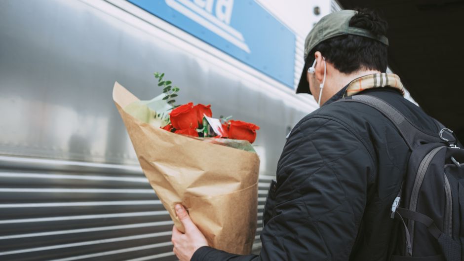 A person with dark hair wearing a beige cap, black jacket, and earphones, holding a bouquet of red roses wrapped in brown paper. The bouquet includes green foliage and is positioned close to their che