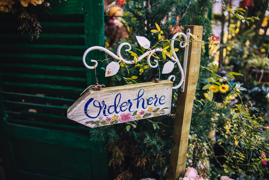 A close-up of a rustic wooden garden sign with the words 'Order here' painted in blue cursive, hanging from a decorative white wrought iron bracket. The sign is surrounded by lush green foliage, inclu