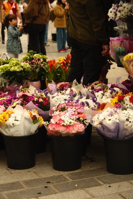 A variety of fresh flower bouquets arranged in black buckets on a busy outdoor street stall, showcasing vibrant colours including pinks, whites, purples, reds, and yellows. The flowers consist of lili