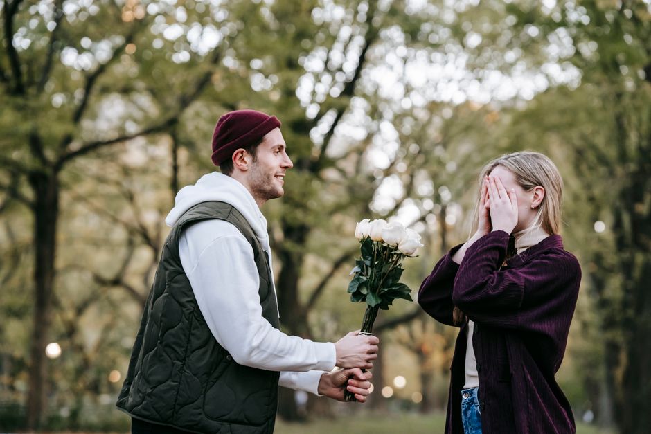 A man dressed in a white hoodie and dark vest, smiling and holding a bouquet of white roses wrapped in dark green foliage, offers the floral gift to a woman with blonde hair wearing a dark purple swea