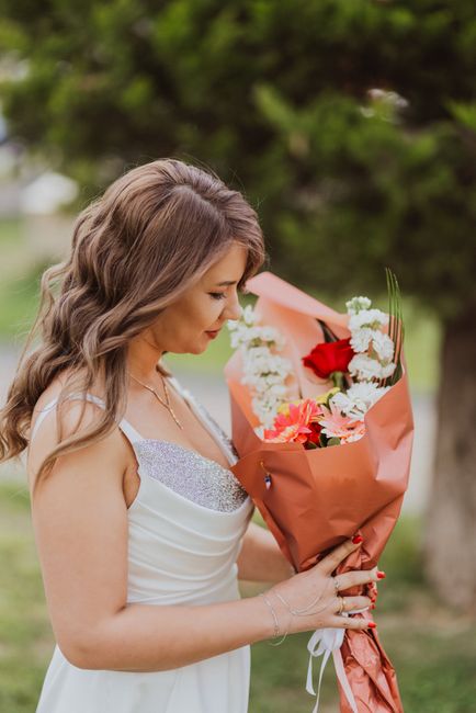 A young woman with long, wavy brown hair is holding a large floral bouquet outdoors against a blurred green leafy background. She is wearing a white sleeveless dress with a sparkly silver detail at th